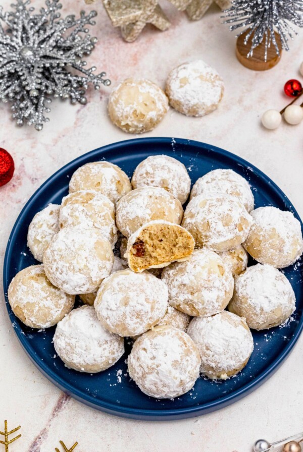 Powder sugar coated Snowball cookies, stacked on a blue plate with holiday party favors on the table around the plate, with one cookie missing a bite.
