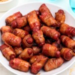 Golden bbq coated bacon wrapped smokies on a white plate with blue glasses of water on the table.