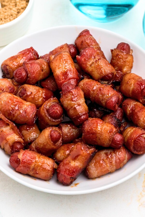 Golden bbq coated bacon wrapped smokies on a white plate with blue glasses of water on the table.