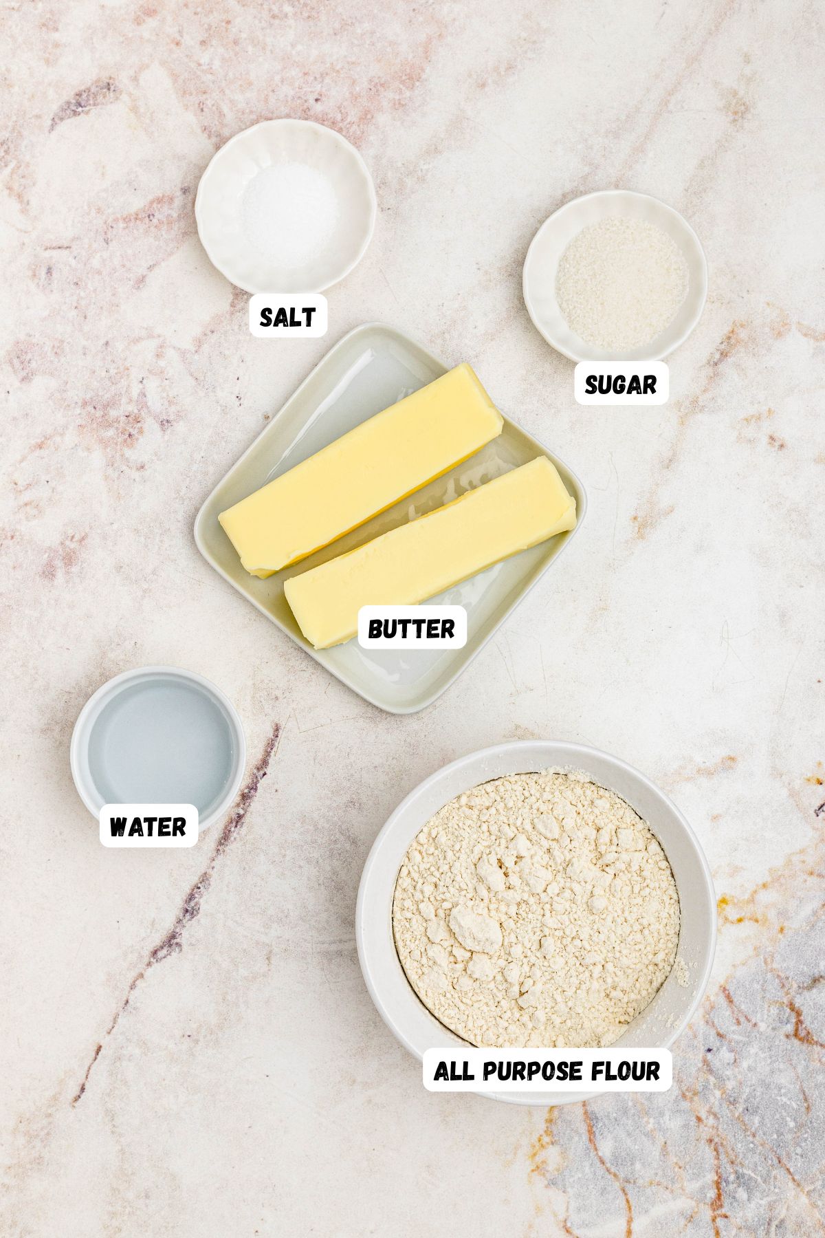 Flour, butter, water, salt, and sugar, measured into small bowls and labeled on a marble table. 