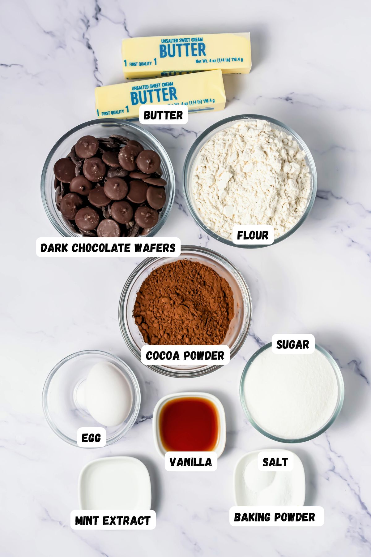 Ingredients like flour, sugar, butter, and more, measured into small bowls on a white marble table and labeled.