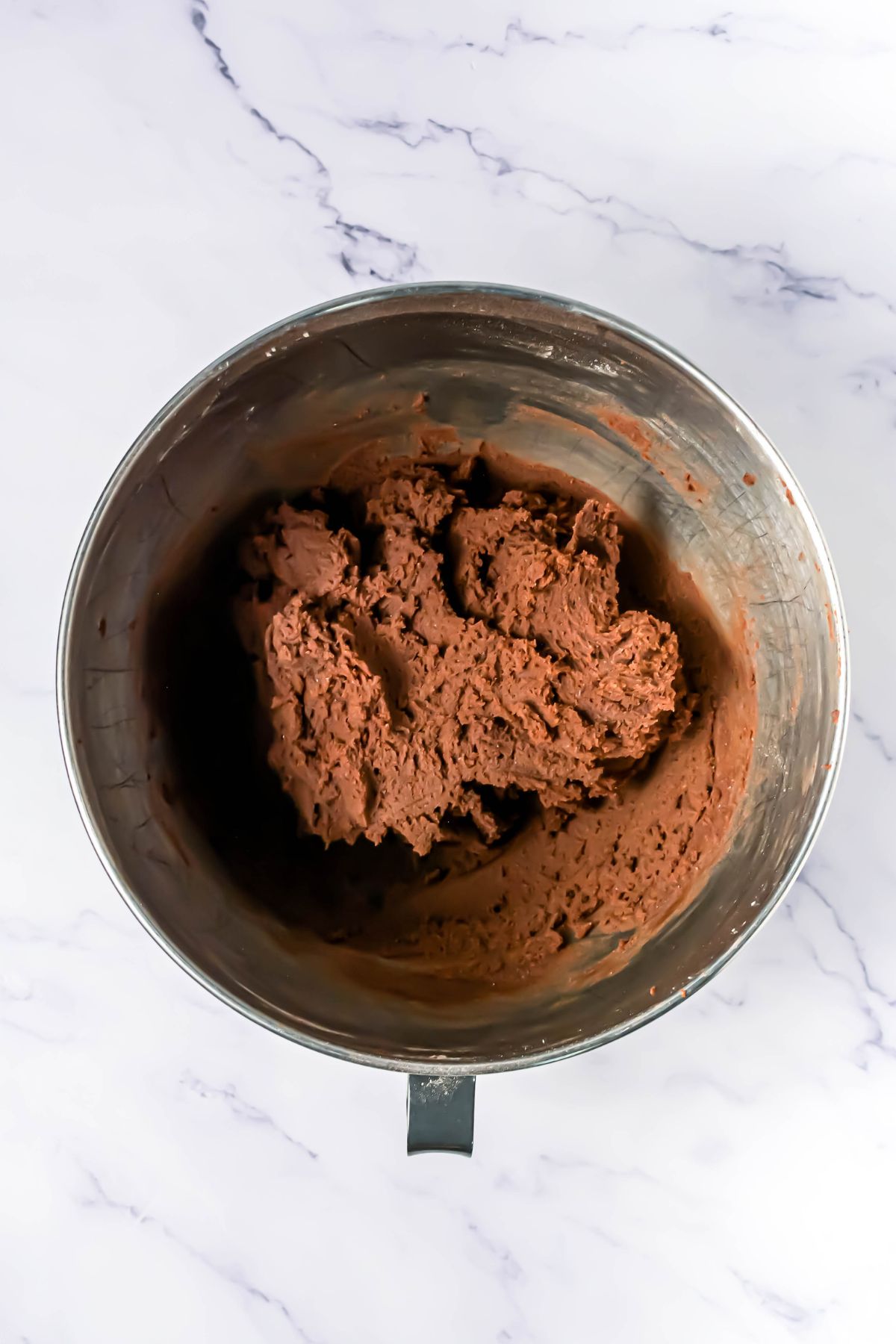 Chocolate cookie dough, formed in a silver mixing bowl, on a white marble table.