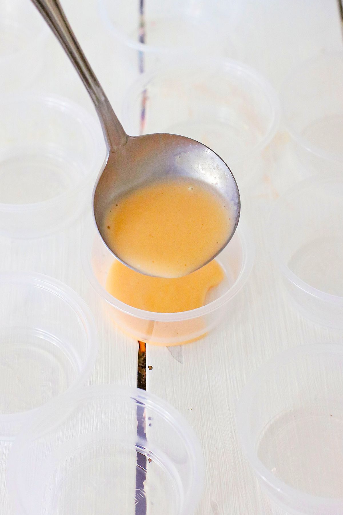 Spoonful of orange mixture being poured into plastic small cups on a white wooden table. 