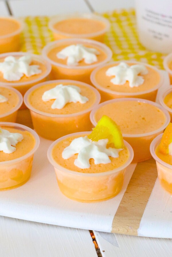 Orange Jello shots in small plastic containers, topped with whipped cream and a small orange wedge, on a wooded table.