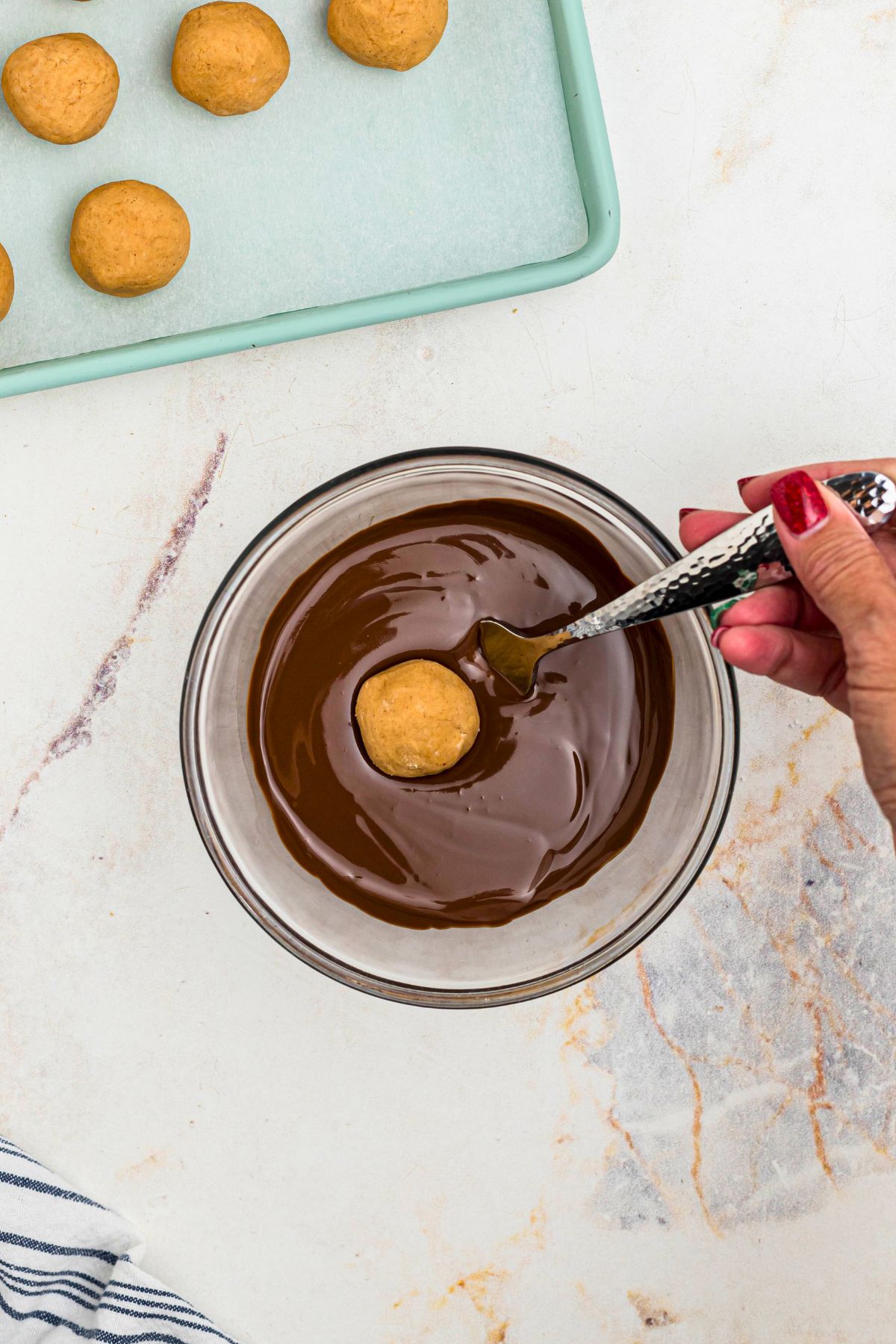 Melted chocolate in a clear glass bowl, with a peanut butter ball being dipped into the bowl. 