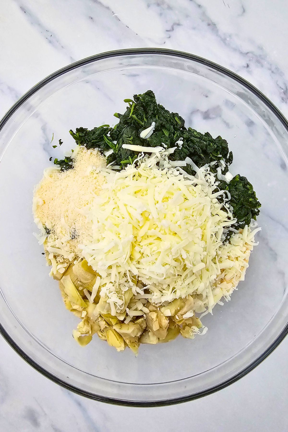 Spinach, artichoke, and cheeses, in a large clear glass mixing bowl. 
