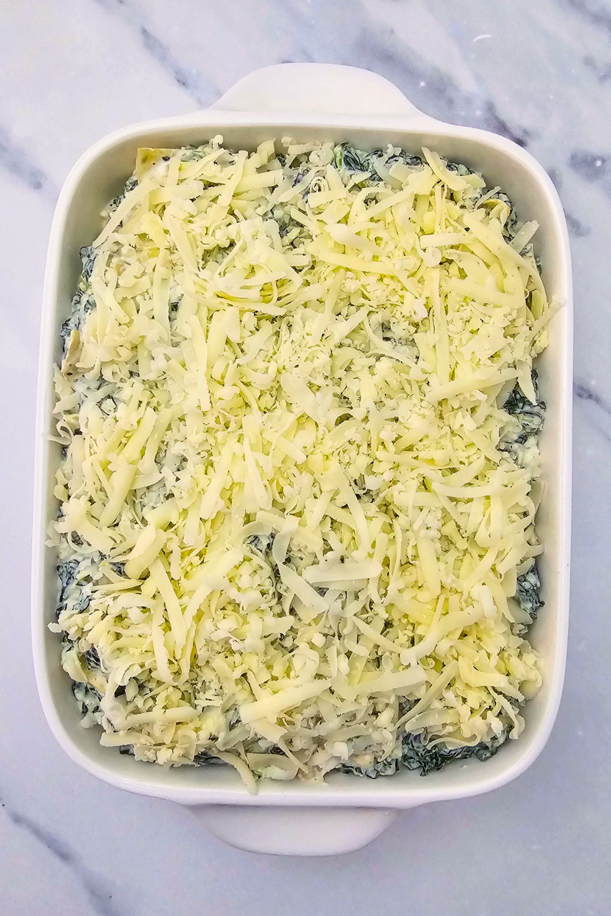 Uncooked cheeses with spinach and artichokes, in a rectangle dish, topped with shredded cheese, on a marble table. 