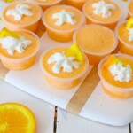 Orange Jello shots in small plastic containers, topped with whipped cream and a small orange wedge, on a wooded table.