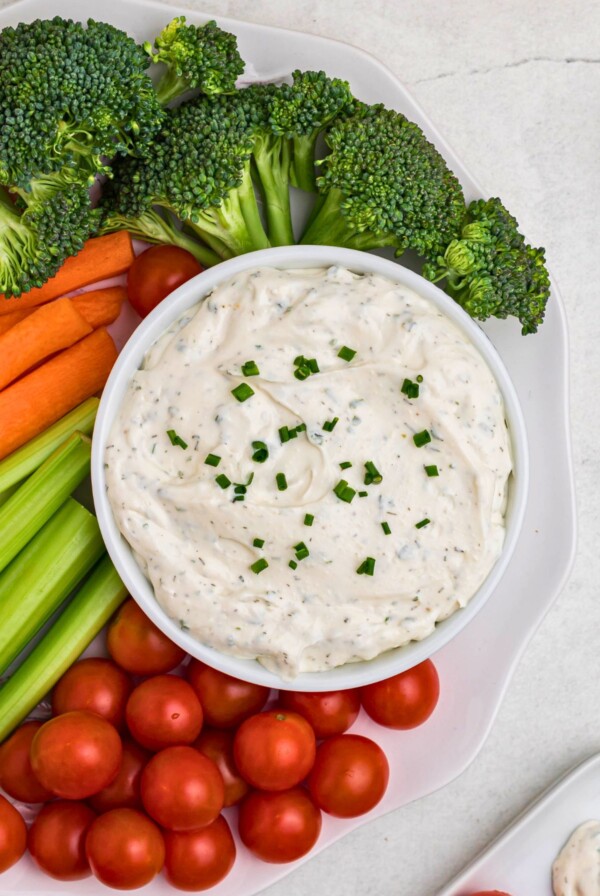Small white bowl filled with dip, surrounded by fresh vegetables on a white plate, on table.