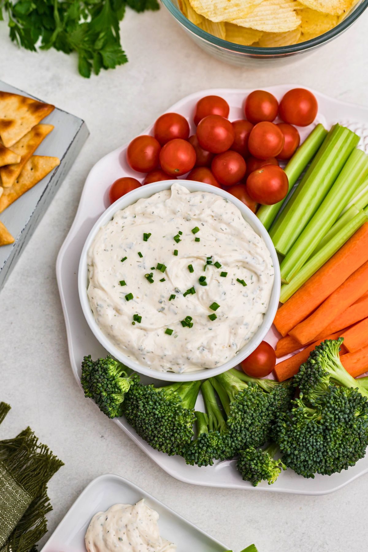 Small white bowl filled with dip, surrounded by fresh vegetables on a white plate, with a plate of potato chips and pita chips also on table.