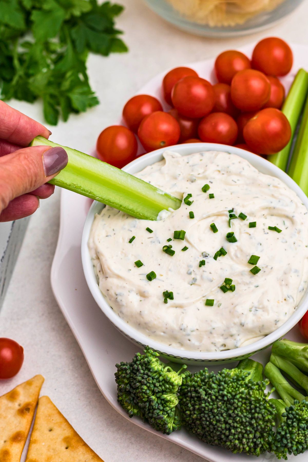 Small white bowl filled with dip, surrounded by fresh vegetables on a white plate, with a plate of potato chips and pita chips also on table. In the center of the image is a hand with a celery stick, dipping out ranch dip.