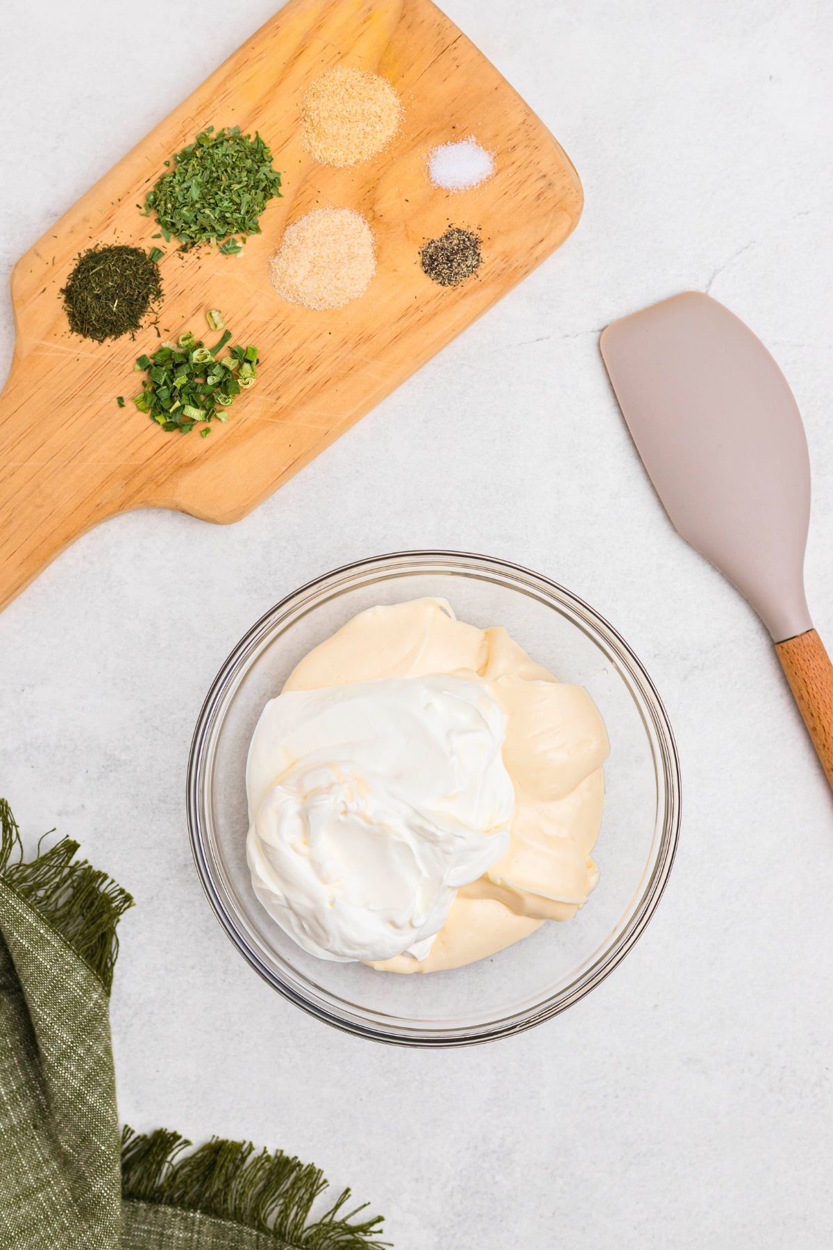 Mayonnaise and sour cream in a clear glass bowl with seasonings and dried herbs on a cutting board next to the bowl, before ingredients are mixed together.
