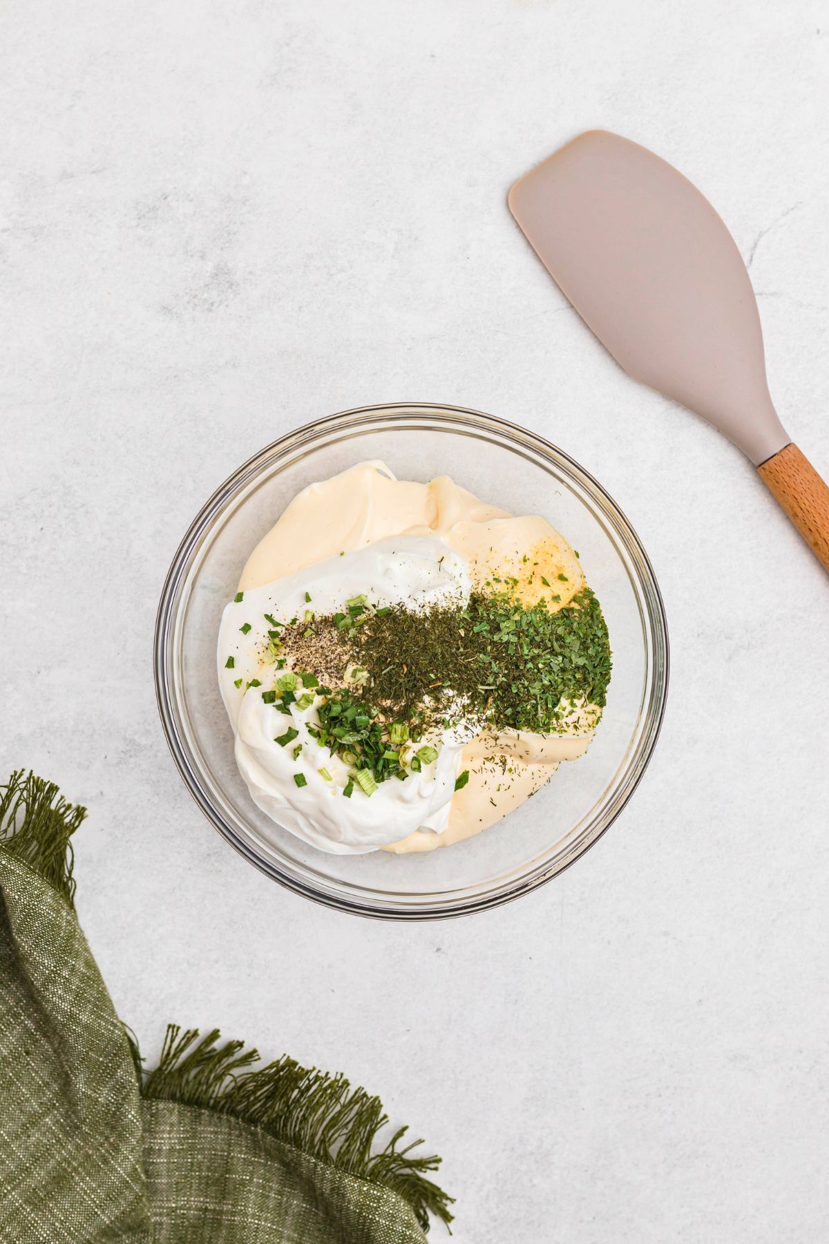 Dried herbs and seasonings added to mayonnaise and sour cream in a clear glass bowl before mixing together to make Ranch dip.