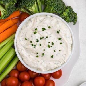 Small white bowl filled with dip, surrounded by fresh vegetables on a white plate, on table.