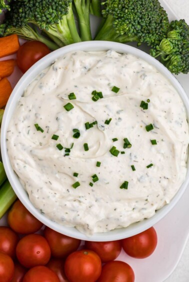 Small white bowl filled with dip, surrounded by fresh vegetables on a white plate, on table.