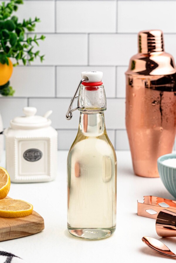 Clear glass bottle of simple syrup on a table, with lemon slices and bar mixing tools next to the bottle.