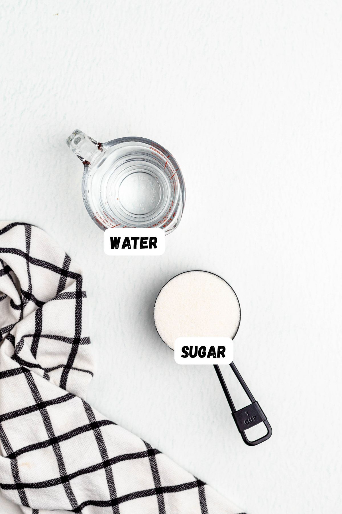 Measuring cup of sugar and measuring cup of water, labeled on a table with a black and white linen. 