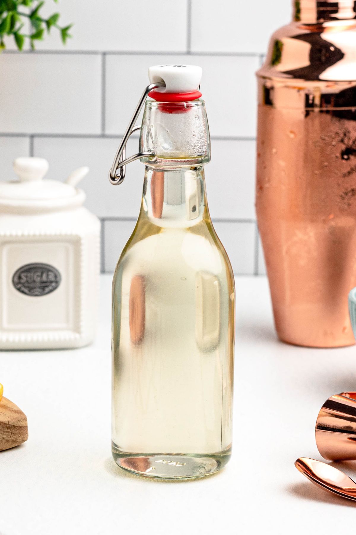 Clear glass bottle of simple syrup on a table, with lemon slices and bar mixing tools next to the bottle. 