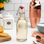 Clear glass bottle of simple syrup on a table, with lemon slices and bar mixing tools next to the bottle.