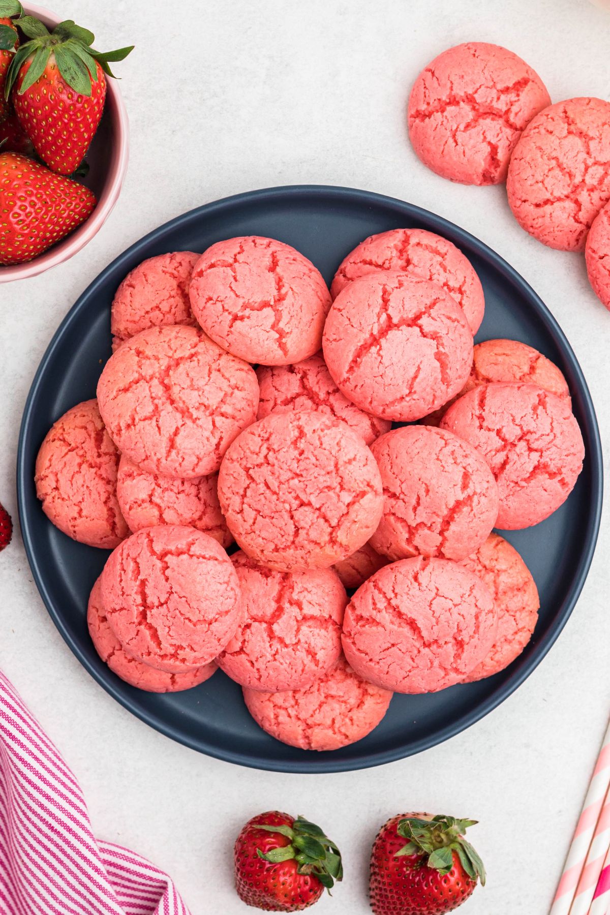 Pink strawberry cake mix cookies stacked on a blue plate in front of glasses of milk and bowls of strawberries.