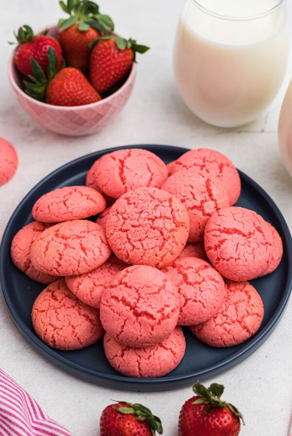 Pink strawberry cake mix cookies stacked on a blue plate in front of glasses of milk and bowls of strawberries.