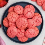 Pink strawberry cake mix cookies stacked on a blue plate in front of bowls of strawberries.