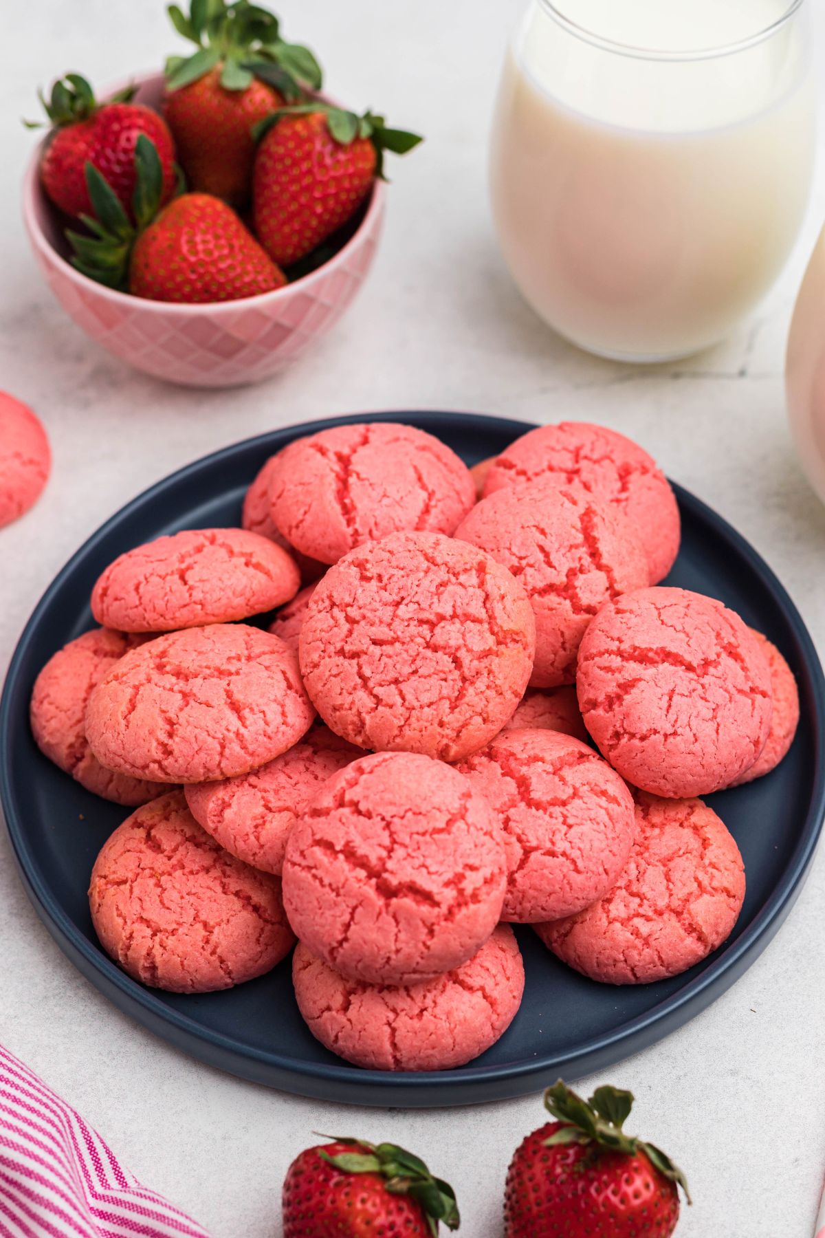 Pink strawberry cake mix cookies stacked on a blue plate in front of glasses of milk and bowls of strawberries.