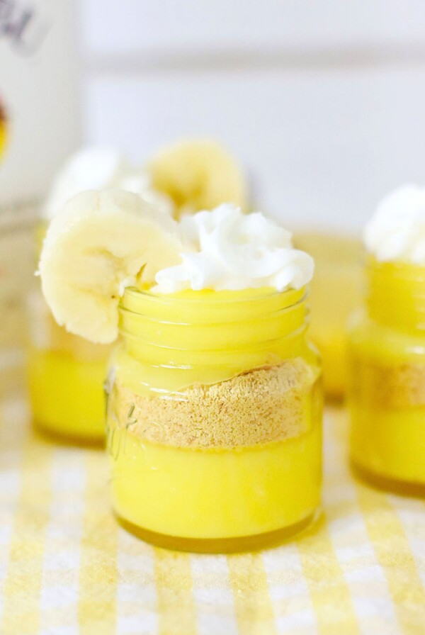 Bright yellow pudding layers in mini jars, topped with whipped cream and garnished with a banana slice.