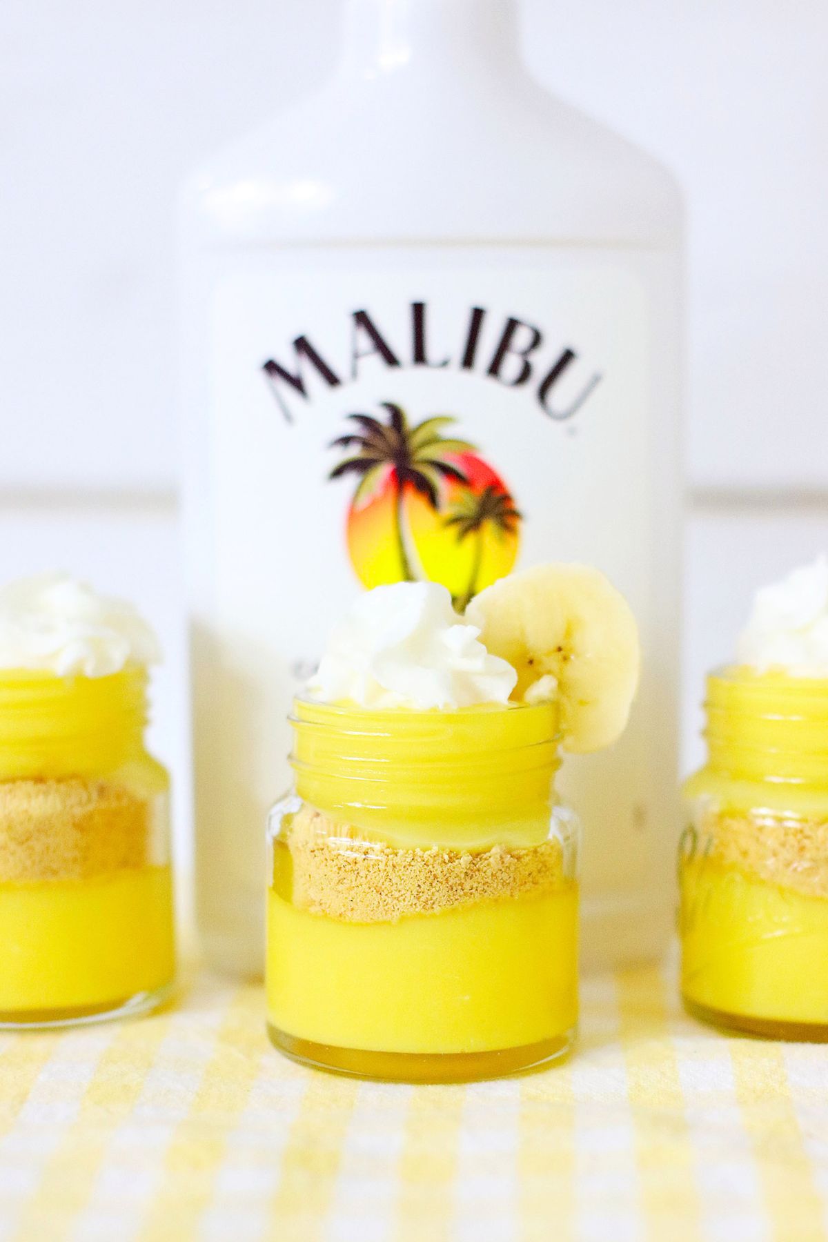 Bright yellow pudding layers in mini jars, topped with whipped cream and garnished with a banana slice in front of a bottle of rum. 
