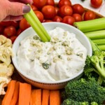 Round white bowl filled with blue cheese dip, surrounded by veggies, with a piece of celery being dipped into center.