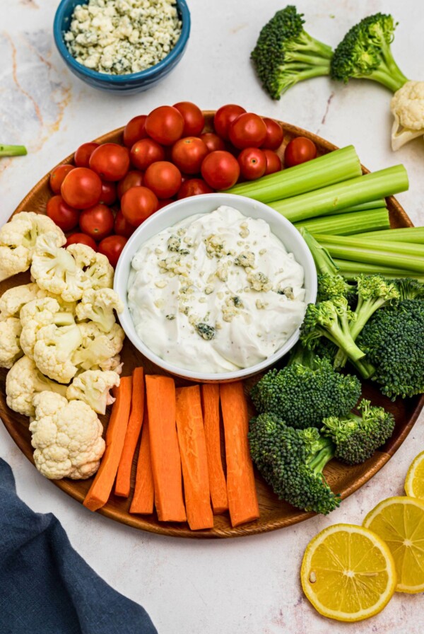 Creamy dip in a small round white bowl, topped with crumbled blue cheese, surrounded by freshly chopped veggies.