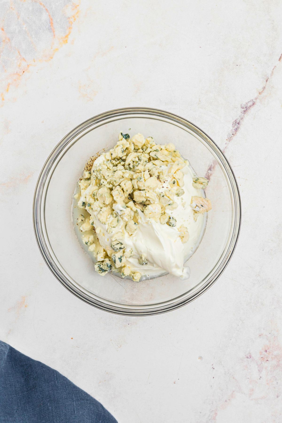 Clear glass bowl with sour cream, blue cheese, and other ingredients, before being mixed together. 