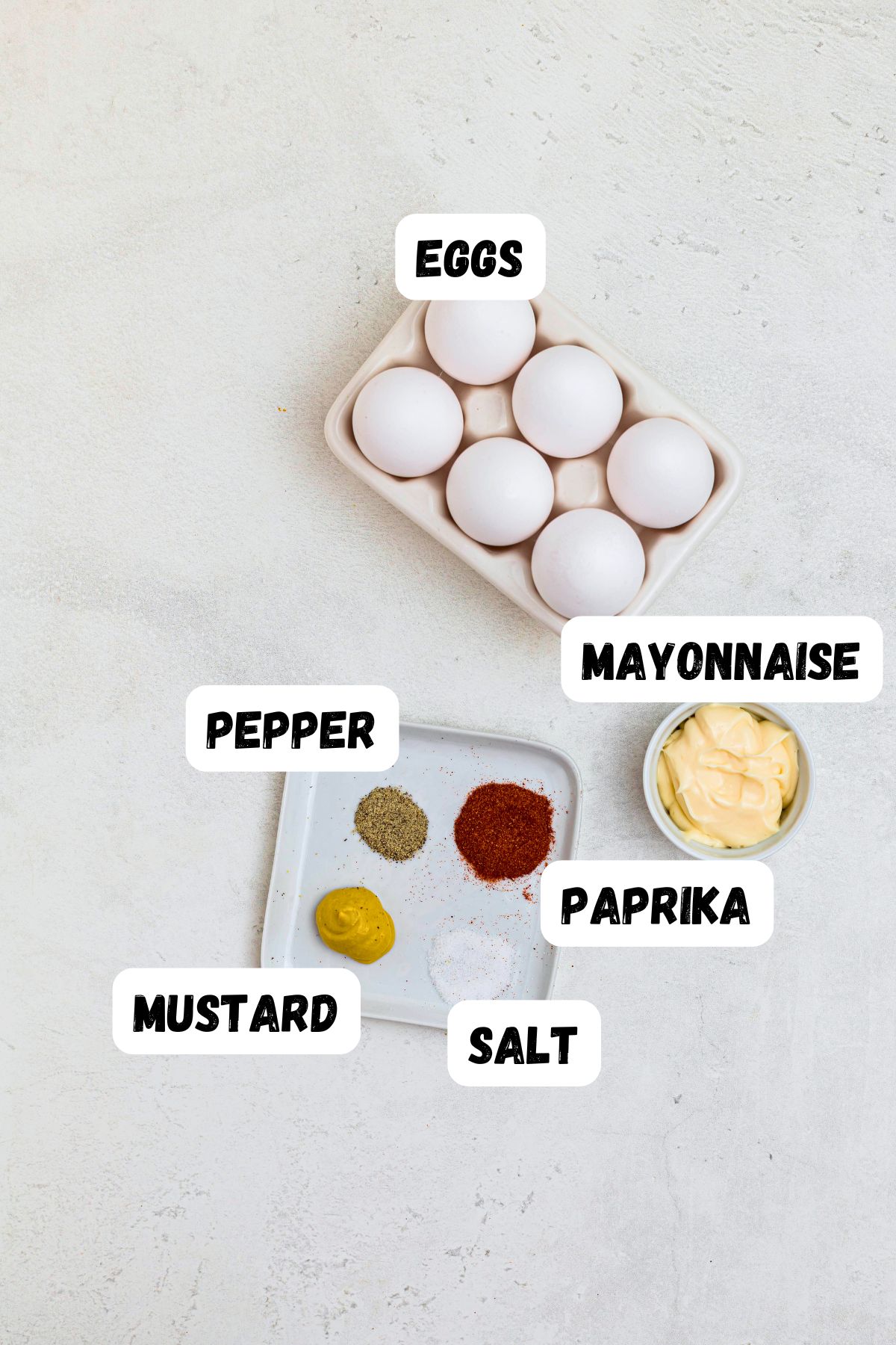 Eggs, mayo, mustard, salt, pepper, and paprika, measured out and labeled on a white marble table.