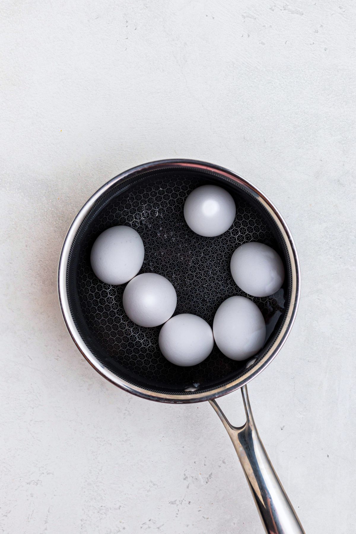 Six whole eggs in shells in a saucepan, covered in water, on a marble table, before boiling. 
