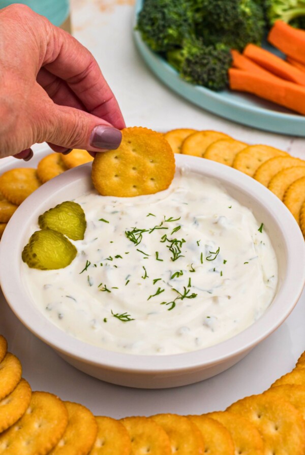 Small white bowl filled with creamy white dip, topped with slices of pickles, surrounded by round crackers, and a plate of fresh vegetables.