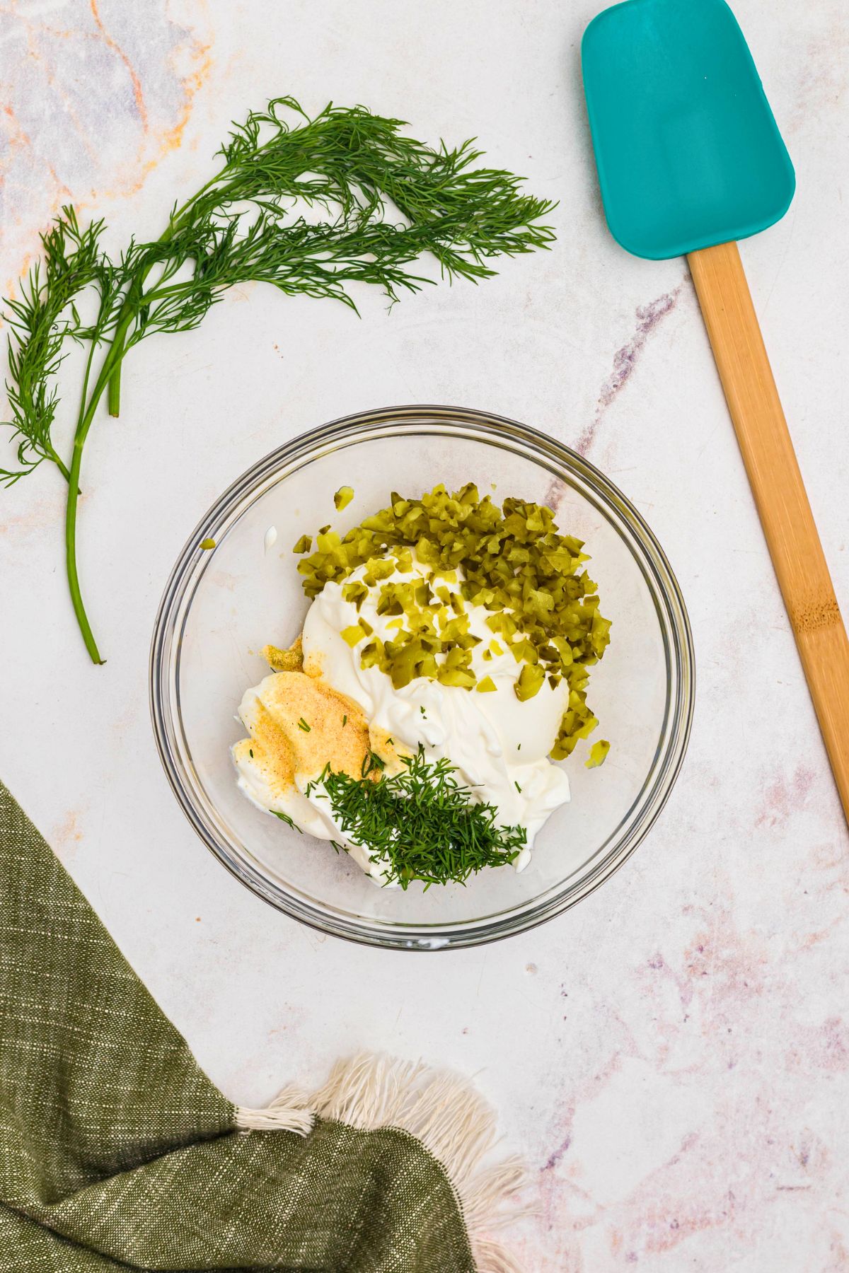 Sour cream, chopped pickles, and other ingredients, in a clear glass bowl, before being mixed together.