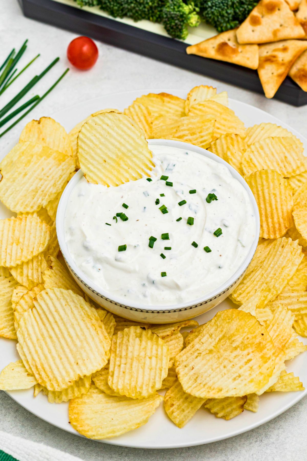 Large plate of potato chips with small round bowl in the center of chips, filled with white creamy sour cream dip, surrounded by fresh vegetables on a platter, with a chip in the center of the dip. 