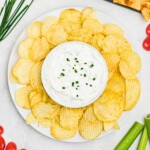 Large plate of potato chips with small round bowl in the center of chips, filled with white creamy dip, surrounded by fresh vegetables on a platter.