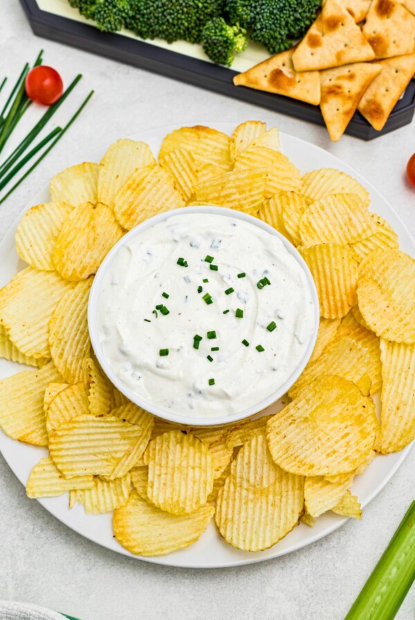 Large plate of potato chips with small round bowl in the center of chips, filled with white creamy dip, surrounded by fresh vegetables on a platter.
