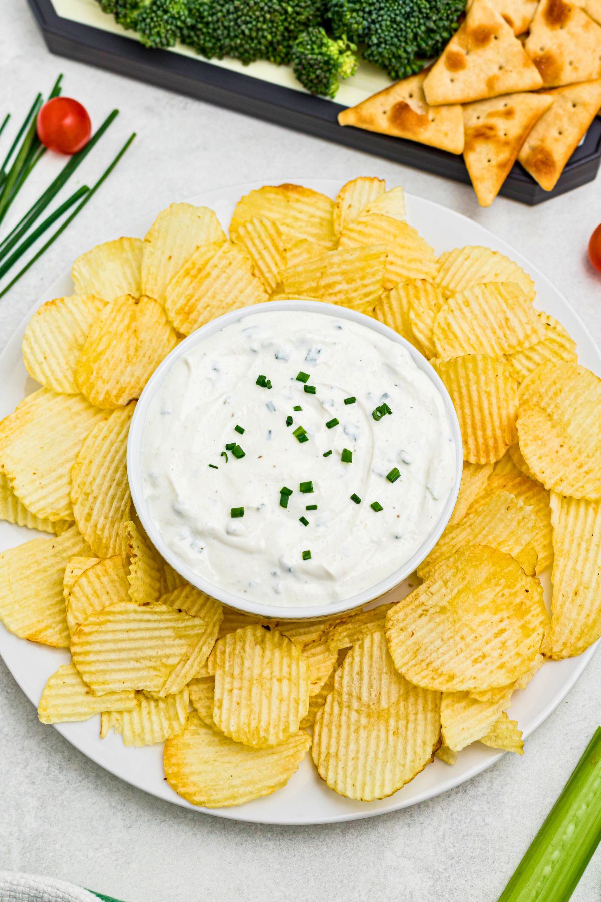 Large plate of potato chips with small round bowl in the center of chips, filled with white creamy sour cream dip, surrounded by fresh vegetables on a platter. 