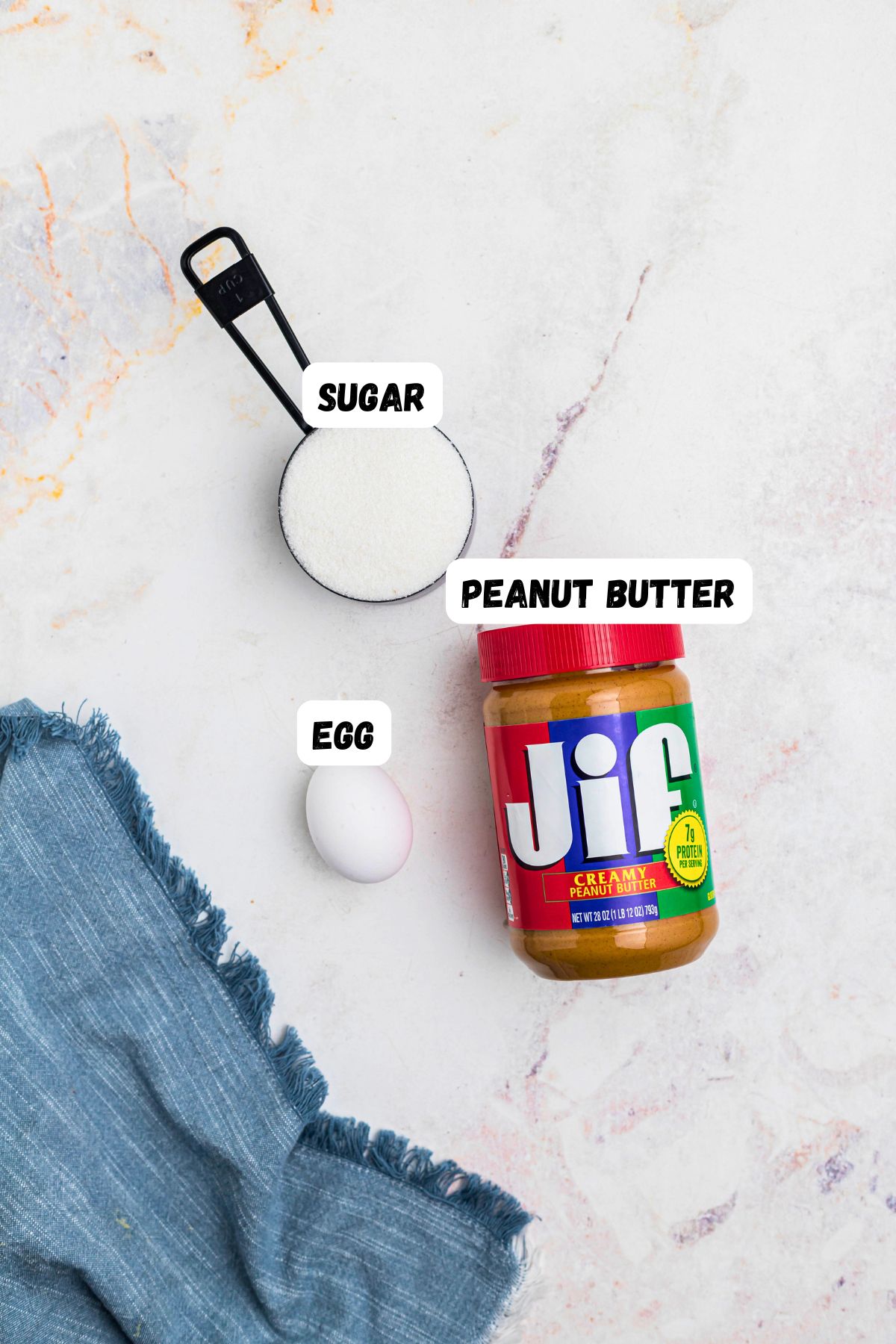 Peanut butter, sugar, and an egg, on a marble table, labeled, next to a blue linen. 
