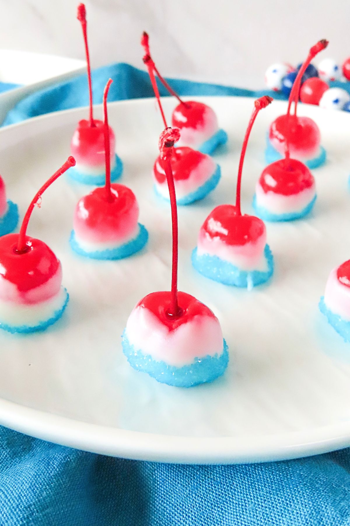 Red cherries, dipped in white chocolate, then dipped again in blue sanding sugar, on a plate waiting to dry. 