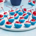 Cherries coated in white chocolate, then dipped in blue sanding sugar, on a white round plate.