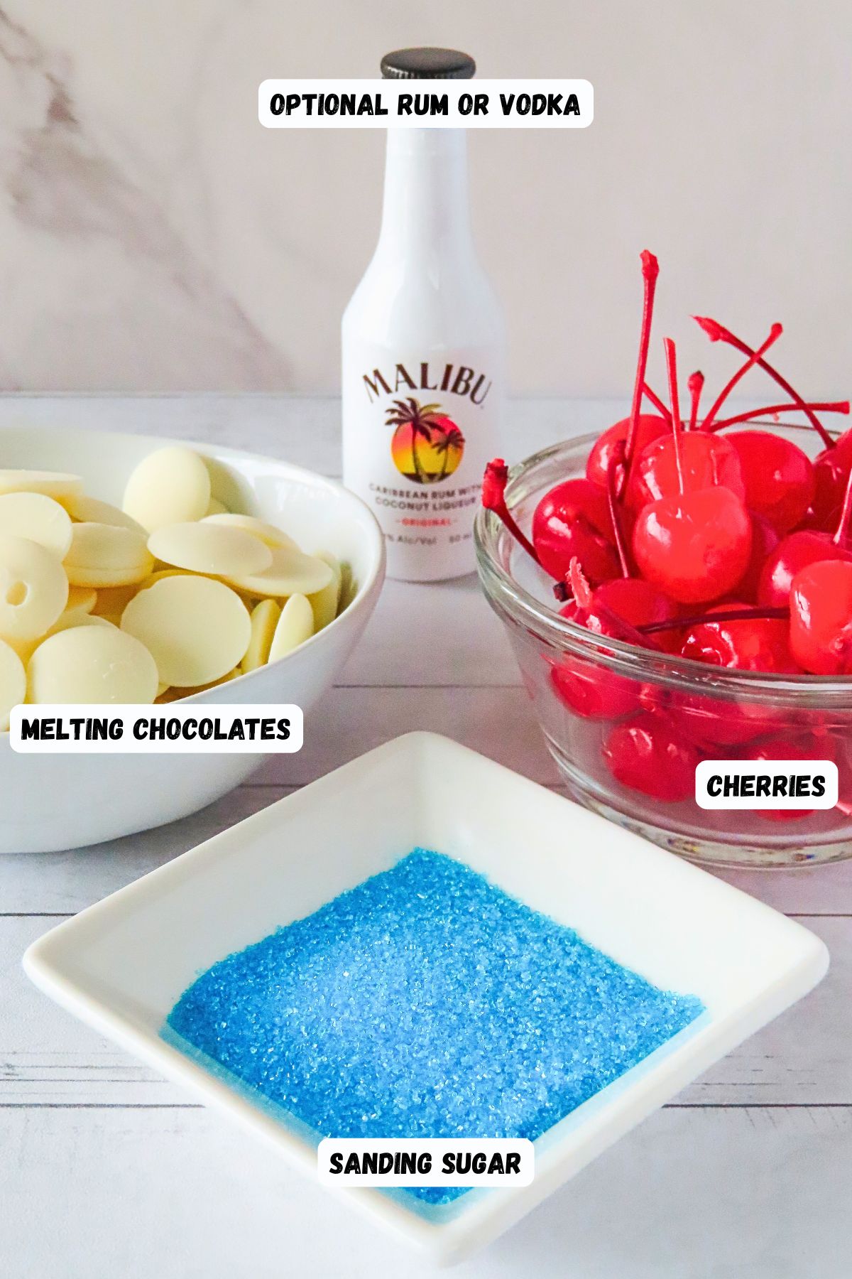 Cherries, white chocolate, sky blue sanding sugar, and optional small bottle of run, on a white wooden tabletop. 