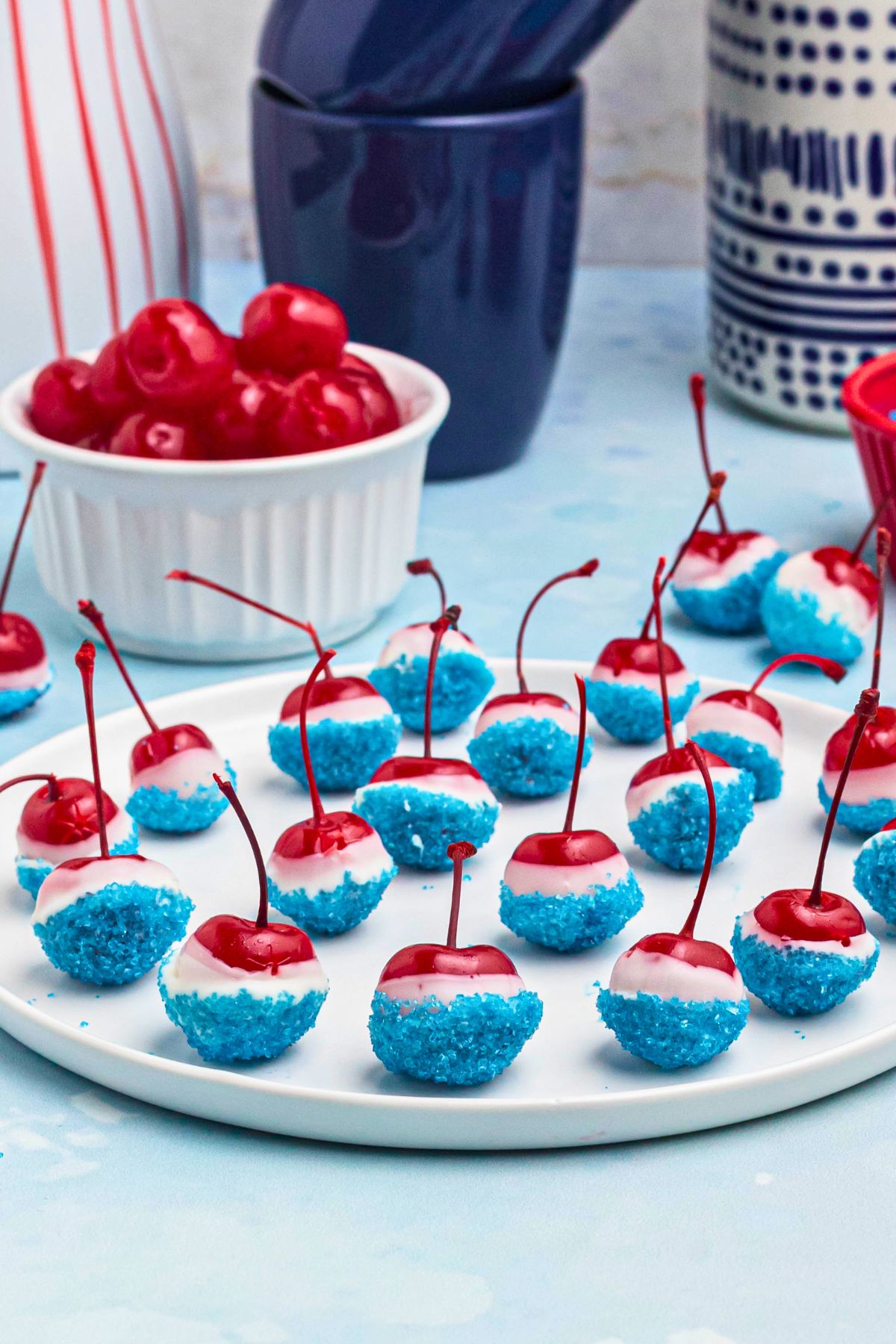 Bright red cherries coated in white chocolate and dipped in blue sanding sugar, on a white plate, with a bowl of cherries and blue coffee mugs in the background.