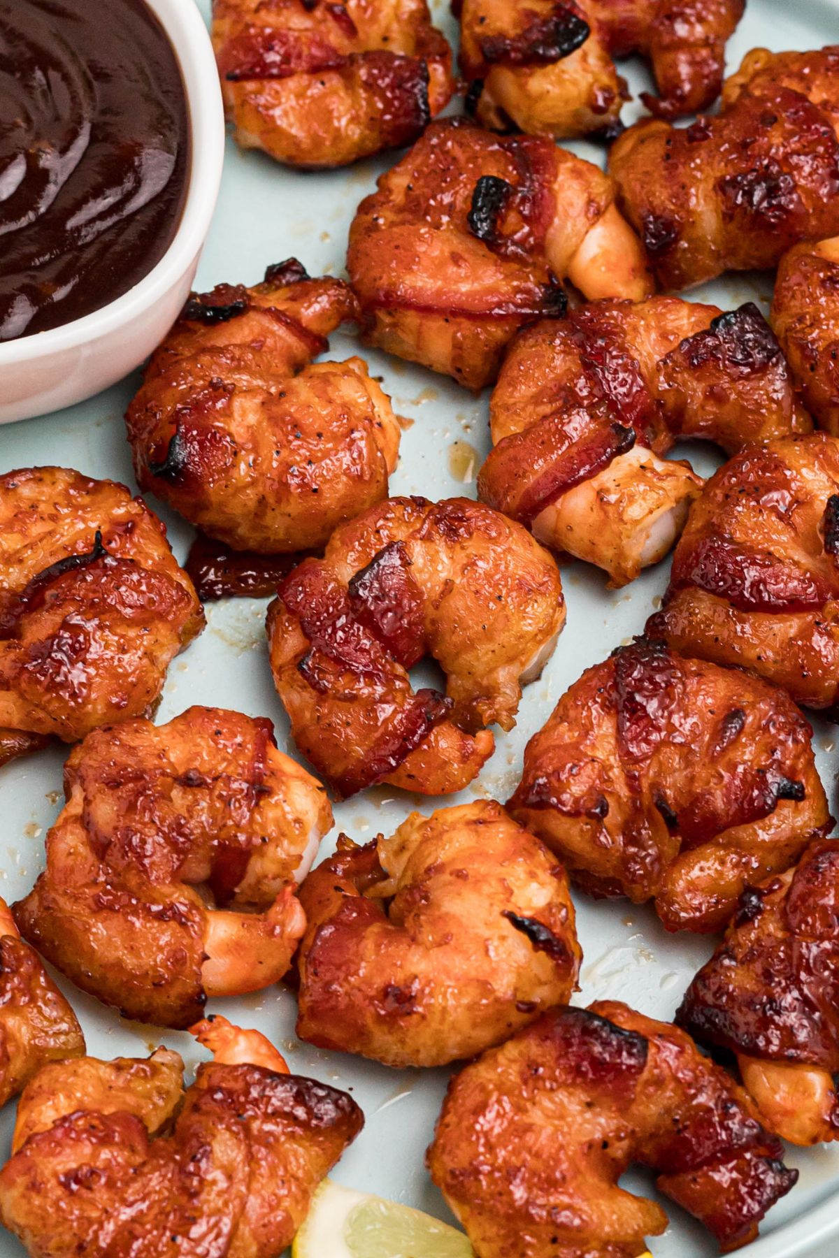 Crispy bacon wrapped around shrimp, coated with BBQ sauce mixture, on a light green plate with lemon slices and serving tongs on the table.