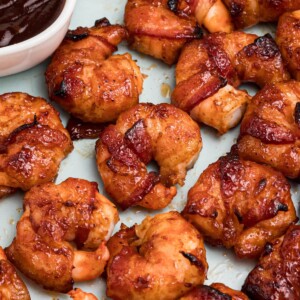 Crispy bacon wrapped around shrimp, coated with BBQ sauce mixture, on a light green plate with lemon slices and serving tongs on the table.