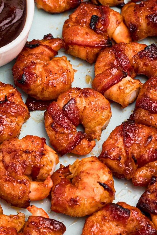 Crispy bacon wrapped around shrimp, coated with BBQ sauce mixture, on a light green plate with lemon slices and serving tongs on the table.