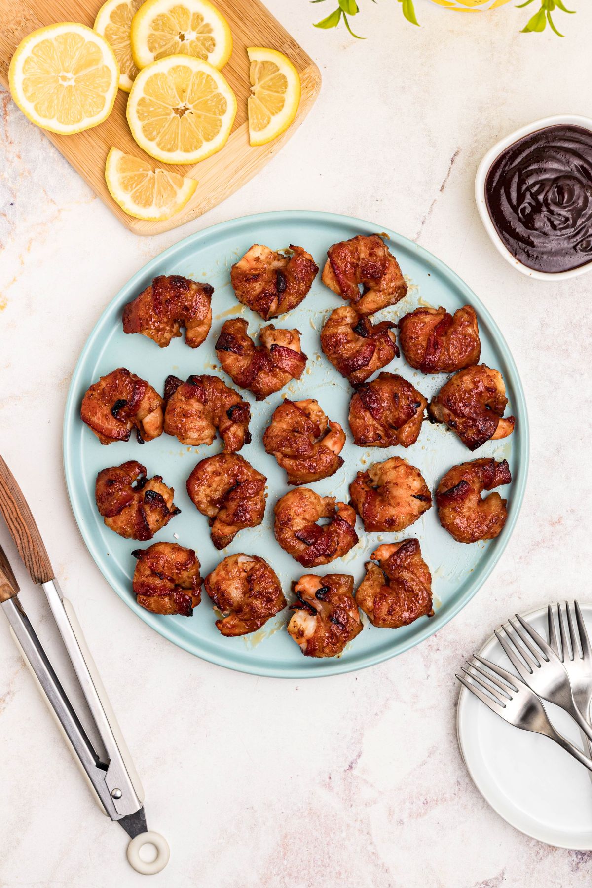 Crispy bacon wrapped around shrimp, coated with BBQ sauce mixture, on a light green plate with lemon slices and serving tongs on the table. 