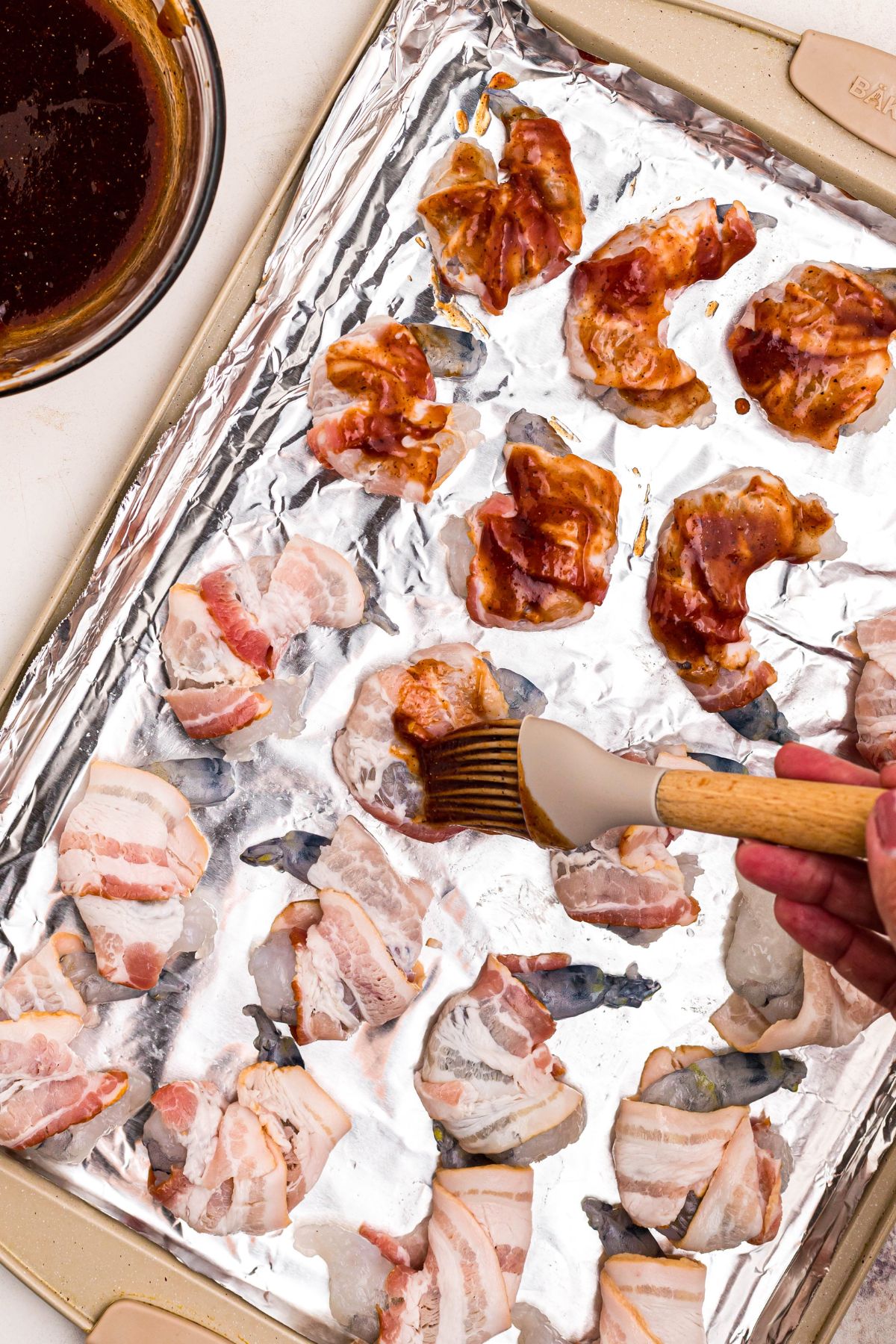Shrimp wrapped in bacon, being brushed with BBQ sauce mixture, on a baking sheet lined with foil. 